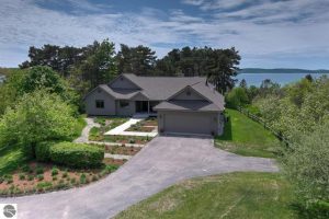Benzie County home for sale with Crytstal Lake shared access and views from an elevated and landscaped lot. This drone photo shows the house, trees, grassy lawn and view of the lake.