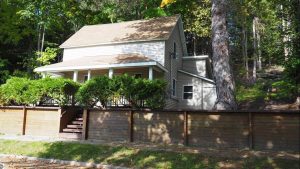 Village of Honor, Benzie County image. Two story farmhouse on a level lot with a wooded hillside behind it. Shade trees and sunny areas, wooden retaining wall with tall bushes behind it lend privacy from the sidewalk