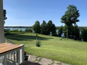 Long Lake Waterfront Home Benzie County image is of a corner of the deck overlooking the grassy backyard with trees on the edge of the ridge with Long Lake views beyond them.