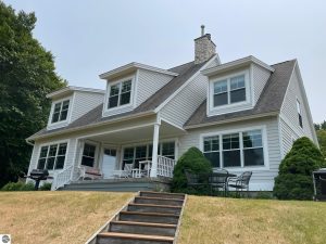 Long Lake Waterfront Home for sale in Benzie County, Image shows the back side of the two story home with numerous windows overlooking the lake and grassy backyard with steps leading to it.