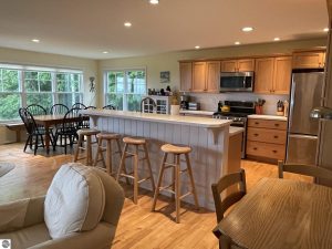 Long Lake Waterfront Home for sale in Benzie County image is looking from the living room toward the kitchen island with stools, the cabinetry, stainless refrigerator and oven, recessed lighting and dining area beyond it.