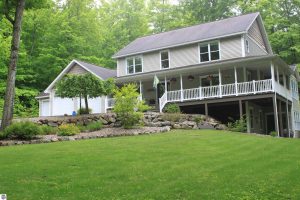 For Sale near Glen Lake image is of a two-story home with a covered wraparound front porch and attached garage on a grassy knoll backdropped by mature trees.