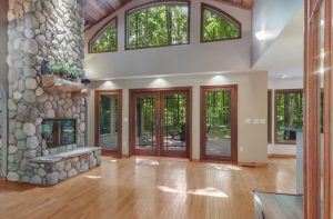 Image 3 of FOR SALE southwest of Lake Ann shows the living room of the home with wood flooring, a cathedral ceiling with upcycled barnwood, eyebrow window and window doors to the back deck with views of the forest and a fieldstone surround fireplace with stone hearth.