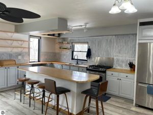 Image for Beulah home on 5 acres is of the snazzy kitchen with an island that has chairs on two sides, open shelves above wooden counters with white cupboards below. A stainless fridge, two windows, overhead lighting and laminate flooring.