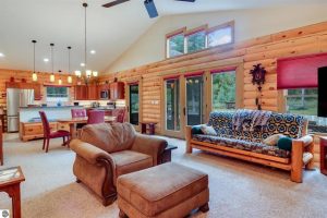 This is a partial view of the carpeted living room with the French doors and windows above them, log-sided wall and kitchen and dining room in the distance. A fireplace is across the room from this scene, out of the frame.