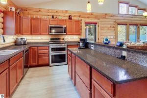 Log home interior photo of kitchen shows the cherry wood cabinetry, stainless gas oven with microwave above it and a cooking island with granite counter. You can see the pine log walls and pendulum lighting. This is a bright and cheery space.