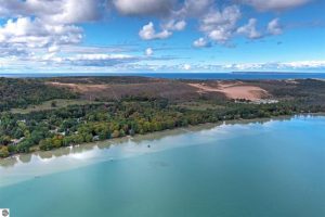 Drone shot is taken over the turquoise water of Little Glen Lake toward the shore with the Dune Climb as a Texas-shaped patch of sand from above and dense forest surrounding it, where the home is, and a sliver of Lake Michigan shows in the distance behind the dunes.