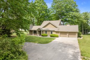 Image for Big Platte Lake Waterfront is a photo of the front of the home facing the driveway. There is a covered front porch, two stall garage and a peak over the main portion of the home. Mature trees surround the grassy yard.