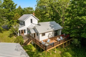 Image for Waterfront Centennial Farm is a close-up of the house taken from a drone clearly show the homes two stories, enclosed front porch and a large deck off the carriage house, with the roof of the garage in the foreground. Mature trees surround the house and the grassy lawn beneath the deck is in sunshine.