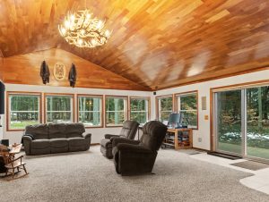 Photo for Smart Cabin Retreat in Benzie County shows the vaulted wood ceiling in the living area with a slider to the outdoors and multiple windows in back. The room is carpeted.