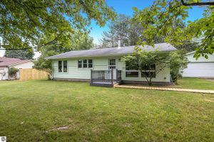 Leelanau Home in Empire photo is of the ranch-style home's front exterior with light-green siding, grassy yard and leafy surroundings. There is a small front deck with sidewalk. Not shown is the breezeway and garage.