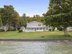 Big Platte Lake home photo is taken from the lake and shows the water, the grassy lawn with a firepit and the home's lake side which includes a deck and 3-season sunroom.